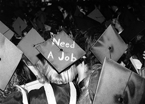 A crowd of several student graduates captured from behind, are dressed in robes and mortarboards.  One mortarboard has written on it “Need a Job”.